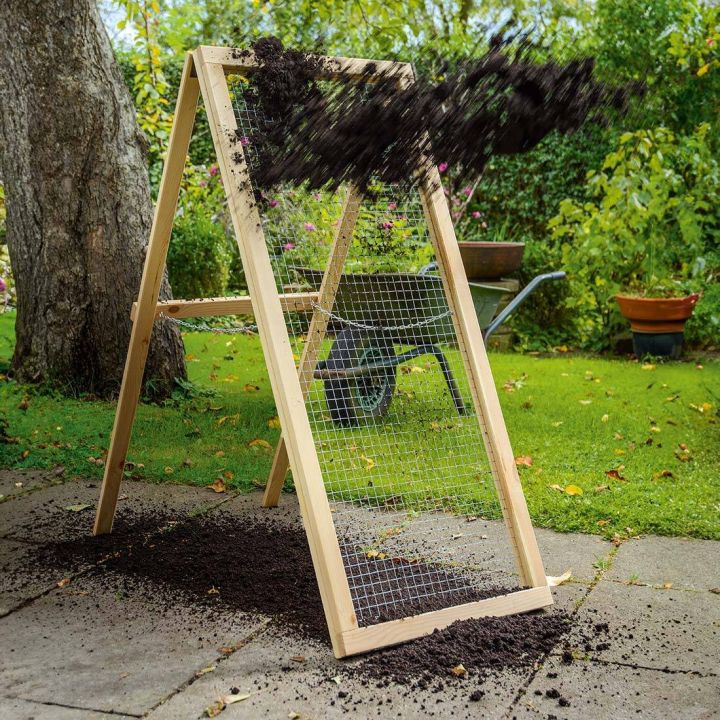 Holzsieb mit Metallgitter in A-Form, stehend auf einer Terrasse, Erde wird darauf gesiebt, Hintergrund: Garten mit Baum, Rasen, Blumen, Schubkarre und Topfpflanze.