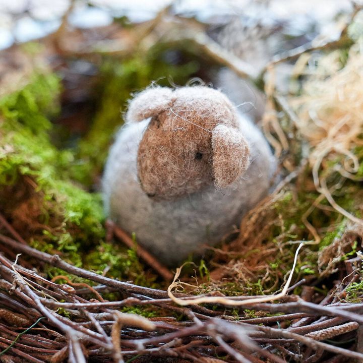 Filzschaf in grauer und brauner Farbe liegt in einem Nest aus Zweigen und Moos.