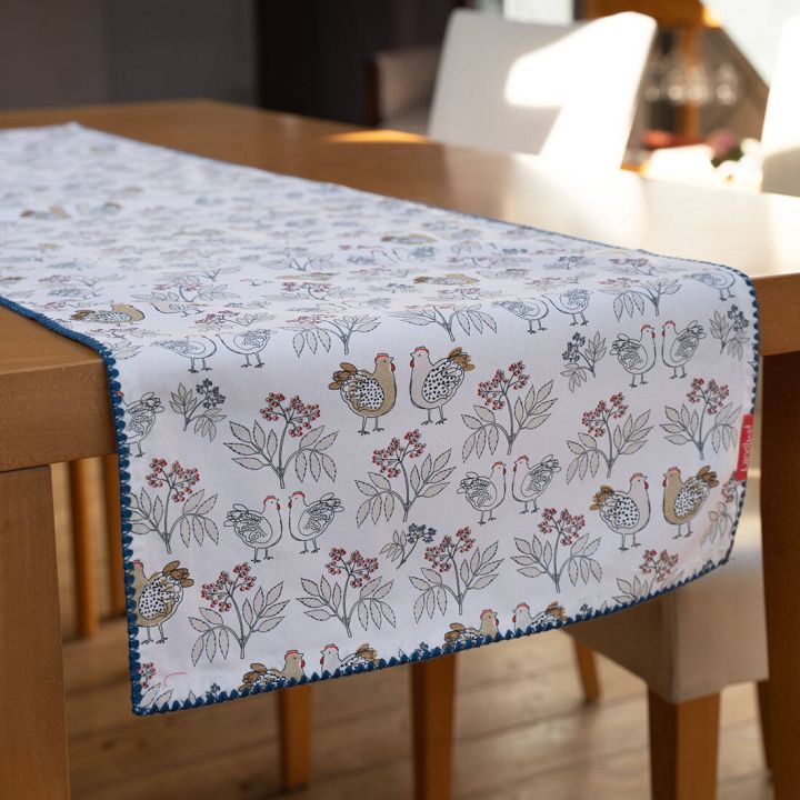 Tischläufer auf Holztisch, weiß mit buntem Hühner- und Blumenmuster, hervorstehende blaue Umrandung.