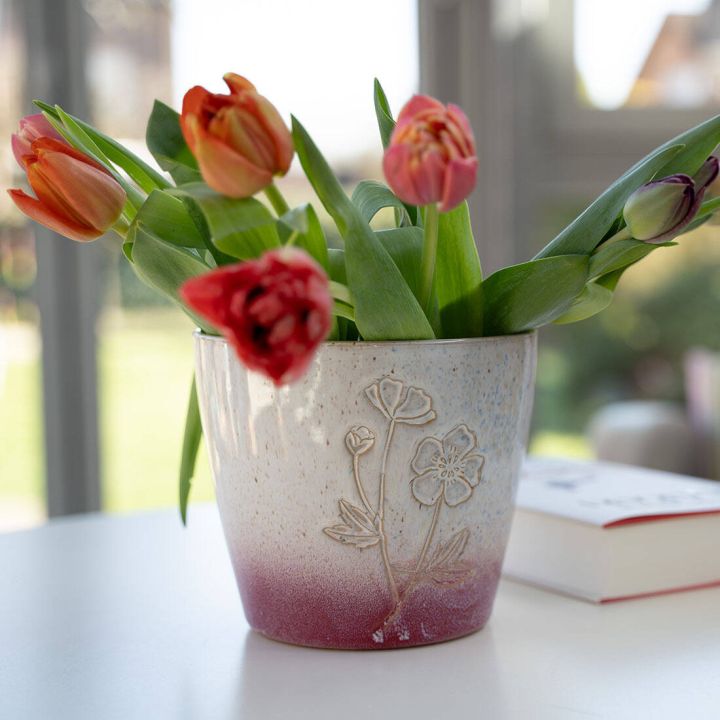 Keramiktopf mit Blumenrelief, gef&uuml;llt mit roten und orangefarbenen Tulpen, steht auf wei&szlig;em Tisch in hellem Raum. Rechts ein geschlossenes Buch.