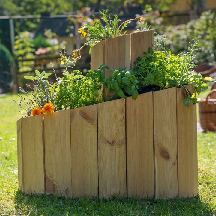Holzpflanzkasten mit drei gestuften Ebenen, bepflanzt mit verschiedenen Kräutern und Blumen, steht auf einer Wiese im Freien. Der Kasten ist aus naturbelassenem Holz gefertigt und zeigt unterschiedliche Pflanzenarten in gestaffelten Höhen.