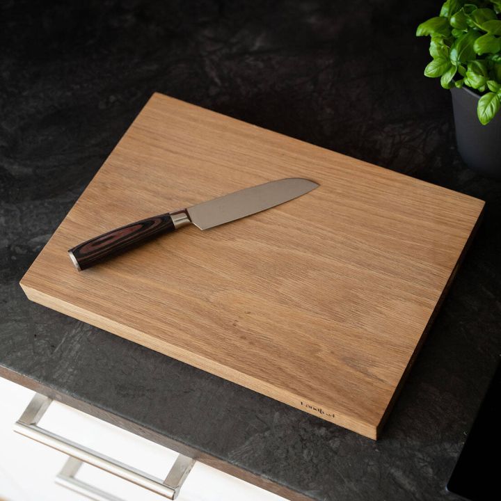 Holzschneidebrett mit Messer auf einer dunklen K&uuml;chenarbeitsplatte. Brett hat seitliche Griffmulden. Rechts befindet sich ein gr&uuml;nes Basilikum in einem Topf.