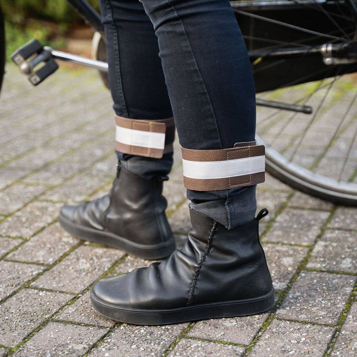 Schwarze Stiefel und enge Jeans mit braun-wei&szlig;en reflektierenden Klettband-Manschetten um die Kn&ouml;chel, stehend neben einem Fahrrad auf gepflastertem Boden.