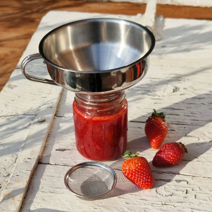 Edelstahltrichter mit Griff sitzt auf einem Glas mit roter Erdbeermarmelade; davor liegen ein feines Metallsieb und drei frische Erdbeeren auf weißem, rustikalem Holztisch.