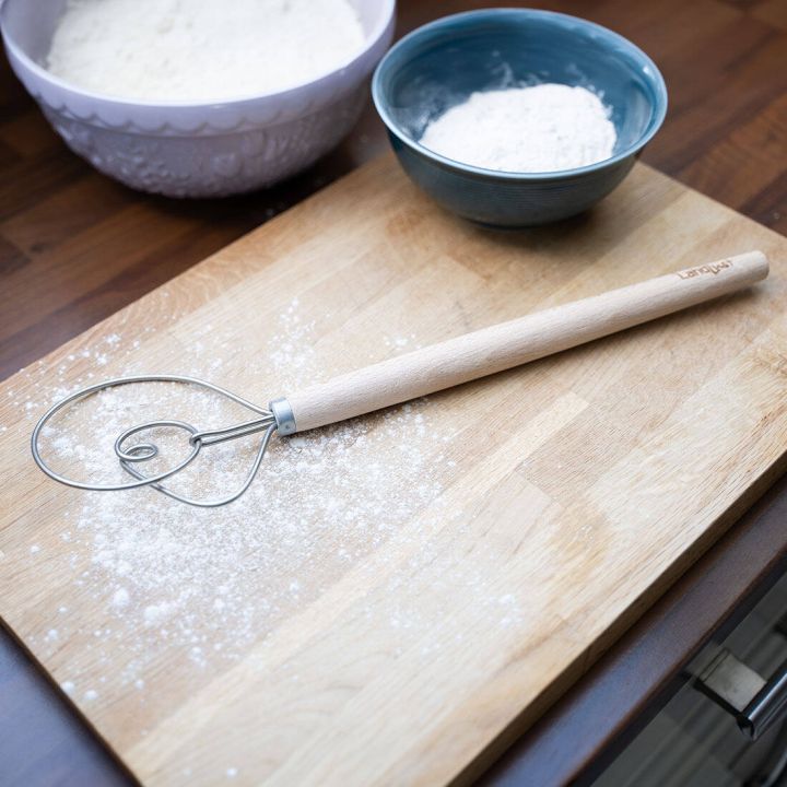 Teigschaber mit Holzgriff und spiralf&ouml;rmigem Edelstahlkopf liegt auf bemehltem Holzbrett; im Hintergrund zwei Sch&uuml;sseln mit Mehl auf einer K&uuml;chenarbeitsplatte.