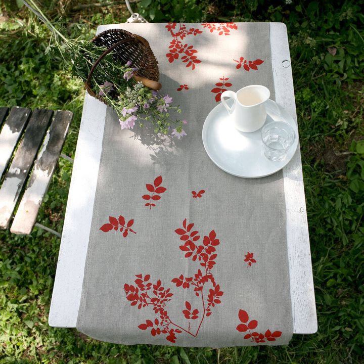 Grauer Tischläufer mit roten Blättermotiven liegt auf einem weißen Holztisch im Freien. Auf dem Tisch stehen eine weiße Kanne, eine Glasschale und ein Korb mit Blumen. Daneben steht eine Holzbank.