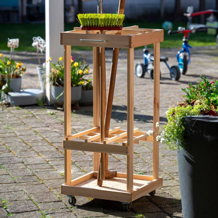 Holzst&auml;nder auf R&auml;dern mit Gartenwerkzeugen, darunter ein Spaten und eine Harke, und einem Pflanzentopf oben. Steht auf einer Terrasse im Freien, umgeben von Blument&ouml;pfen und Kinderspielzeug im Hintergrund.
