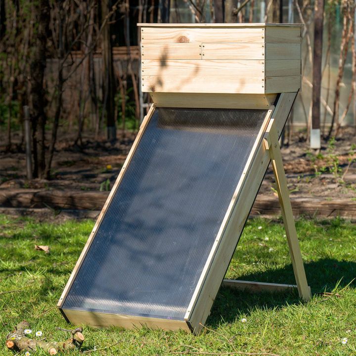 Holzgestell mit geneigtem Solarpanel im Garten aufgestellt. Im Hintergrund sind Bäume und Büsche zu sehen. Das Solarpanel ist schwarz und wird von Holzrahmen gestützt.