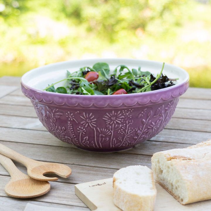 Lila verzierte Schüssel mit Salat, darunter Kirschtomaten und grüne Blätter, auf Holztisch im Freien. Daneben Holzlöffel und angeschnittenes Baguette auf Holzbrett. Unscharfer, grüner Hintergrund.