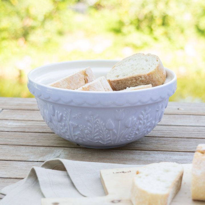 Weiße verzierte Schüssel mit floralen Mustern, gefüllt mit Brotscheiben, auf Holztisch im Freien. Daneben ein Holzbrett mit weiterer Brotscheibe. Unscharfer, grüner Hintergrund.