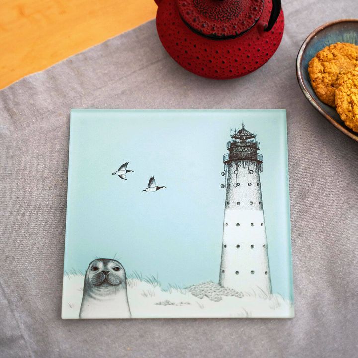 Ein quadratisches Glasbild zeigt einen Leuchtturm, zwei fliegende Vögel und eine lächelnde Robbe im Gras. Es liegt auf einer grauen Tischdecke neben einer roten Teekanne und einem Teller mit Keksen.