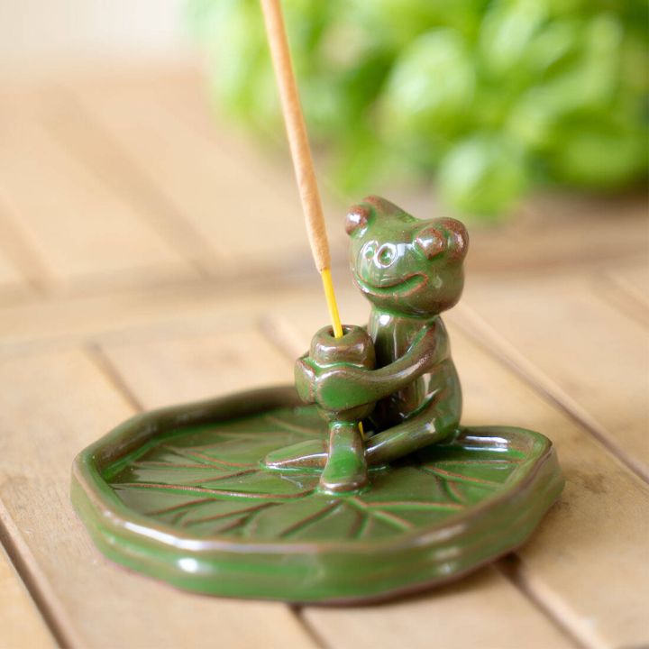 Grüner Keramik-Räucherstäbchenhalter in Froschform, sitzend auf einem Seerosenblatt-Design. Der Frosch hält ein Räucherstäbchen vertikal. Hintergrund verschwommen, Fokus auf dem Halter.