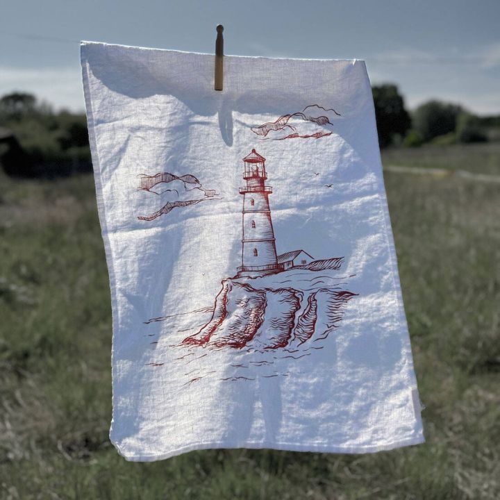 Weißes Geschirrtuch mit roter Zeichnung eines Leuchtturms auf Klippen, aufgehängt an einer Wäscheklammer im Freien. Hintergrund zeigt eine Landschaft bei blauem Himmel.