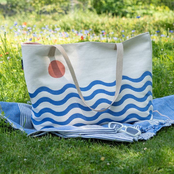 Große Stofftasche mit blauen Wellen und rotem Kreis darauf, steht auf einer blauen Decke, die auf Gras liegt. Eine Sonnenbrille ist auf der Decke platziert. Im Hintergrund ist eine blühende Wiese zu sehen.