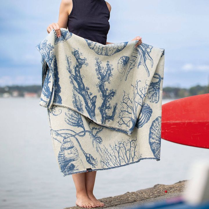 Person hält eine beige Decke mit blauem Meeresmotiv aus Korallen und Muscheln, steht barfuß auf einem Betonboden am Wasser. Die Decke ist detailliert bedruckt, im Hintergrund sichtbar verschwommene Landschaft und rotes Kanu.