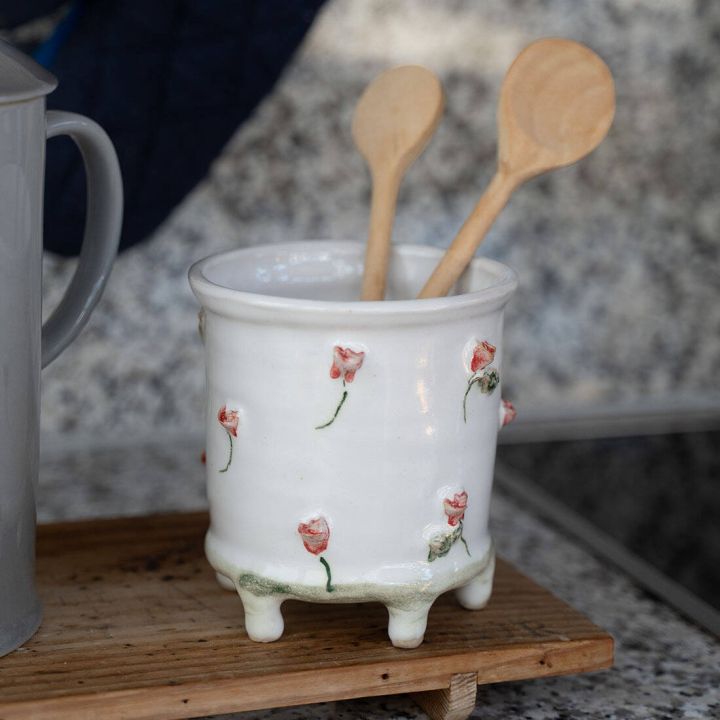 Weißer Keramikbehälter mit kleinen rosa Blumenmotiven steht auf einem Holzbrett. Zwei hölzerne Kochlöffel stecken darin, im Hintergrund ist verschwommenes, granitartiges Muster sichtbar.