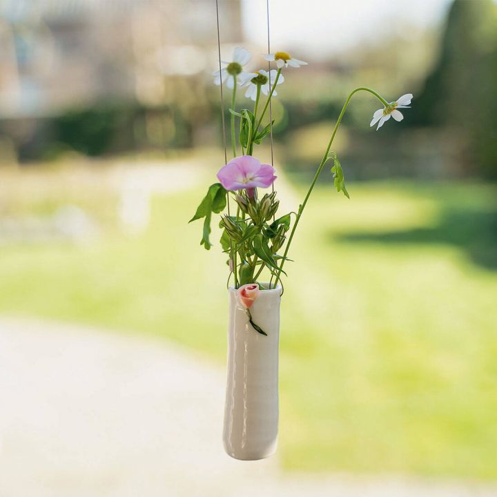 Kleine hängende Vase aus weißer Keramik mit einem rosa Blumenmuster, gefüllt mit Wiesenblumen. Sie hängt an dünnen Fäden, verschwommener grüner Garten im Hintergrund.