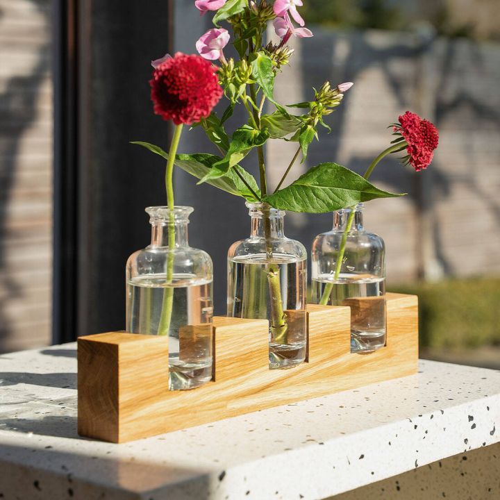 Drei kleine Glasvasen in einem hölzernen Halter, jede mit Wasser gefüllt. Zwei Vasen haben rote Blumen, die mittlere hat rosa Blüten und grüne Blätter. Steht auf einer hellen, gesprenkelten Oberfläche im Freien.