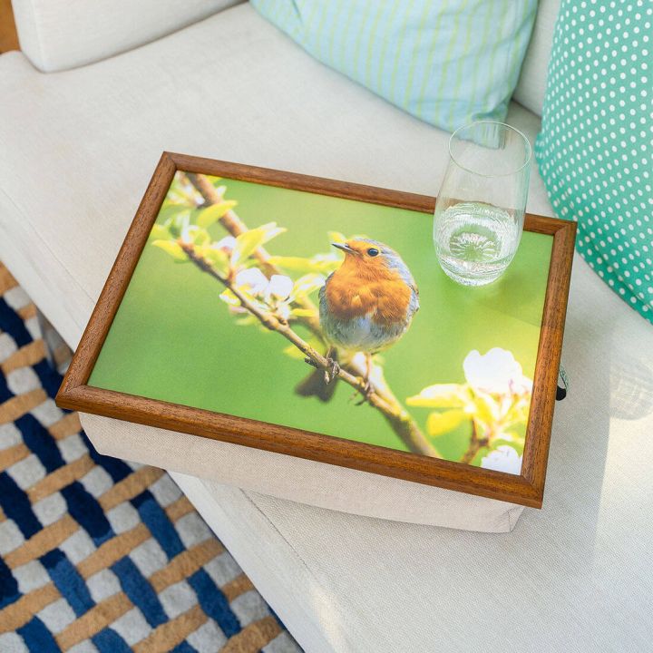 Ein Scho&szlig;tablett mit Holzrahmen und einem Bild eines Rotkehlchens auf einem Zweig. Auf dem Tablett steht ein Glas Wasser. Das Tablett liegt auf einem hellen Sofa mit gemusterten Kissen.