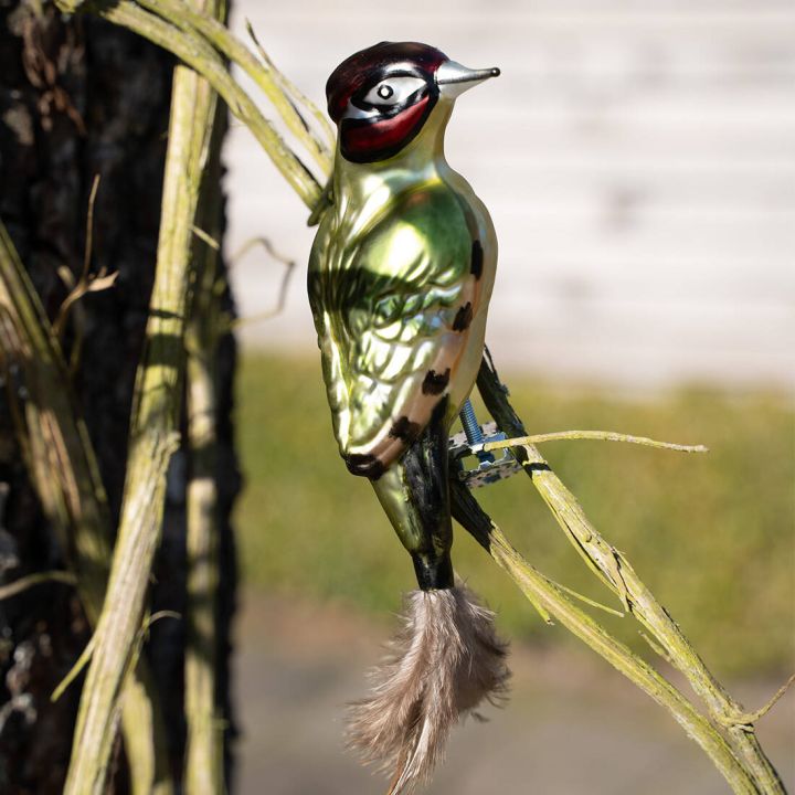 Glasornament in Form eines gr&uuml;n-bunten Vogels mit rotem Kopf, auf einem Ast befestigt, mit Federschwanz, vor unscharfem Hintergrund.