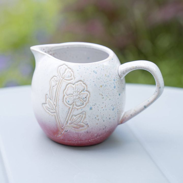 Wei&szlig;er Keramik-Milchkrug mit floraler Gravur und roten Sprenkeln. Hat einen ergonomischen Henkel und eine Ausgusst&uuml;lle. Steht auf einem grauen Tisch mit gr&uuml;nem, unscharfem Hintergrund.