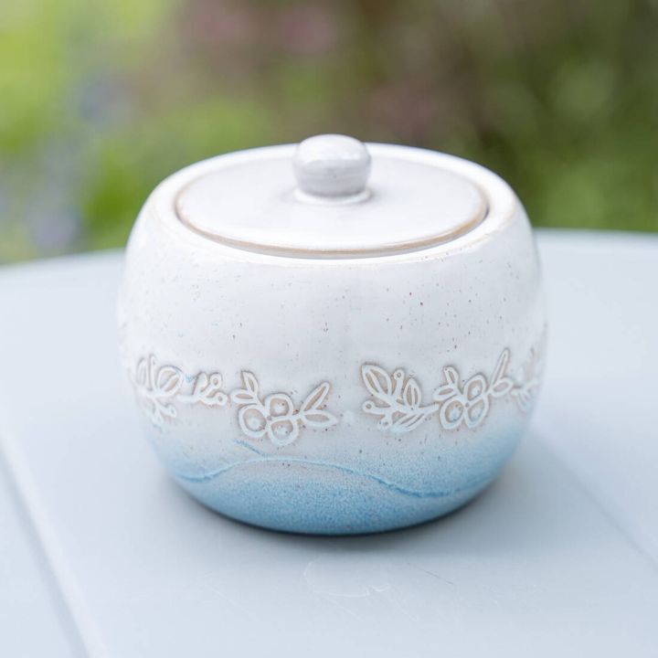 Weißer Keramik-Zuckerdose mit blauem Farbverlauf und floraler Gravur. Mit Deckel versehen. Steht auf einem grauen Tisch mit einem unscharfen, grünen Hintergrund.