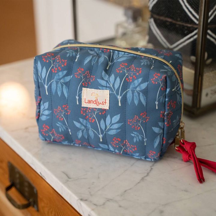 Eine blaue Kosmetiktasche mit Blumenmuster und rotem Beerenmotiv liegt auf einem weiß-marmorierten Tisch. Die Tasche hat einen goldfarbenen Reißverschluss und ein rotes Stoffband als Anhänger. Ein „Landlust“-Label ist sichtbar.