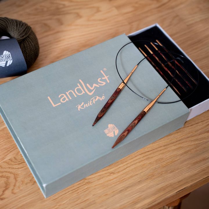 Ein Set von Rundstricknadeln auf einer geöffneten „Landlust KnitPro“-Box liegt auf einem Holztisch. Daneben befindet sich ein dunkelgrünes Wollknäuel.