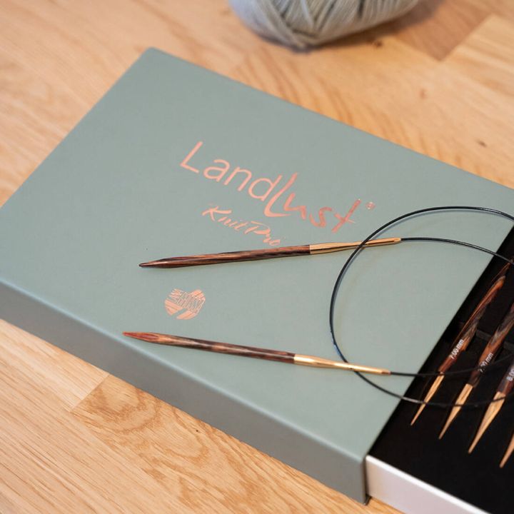 Rechteckige, hellgr&uuml;ne Kartonverpackung mit goldener Aufschrift &bdquo;LandLust KnitPro&ldquo; steht auf einem Holzuntergrund. Vor der Schachtel liegt eine Rundstricknadel, rechts am Bildrand ist ein Kn&auml;uel hellgraues Garn zu sehen. Der Hintergrund ist neutral.