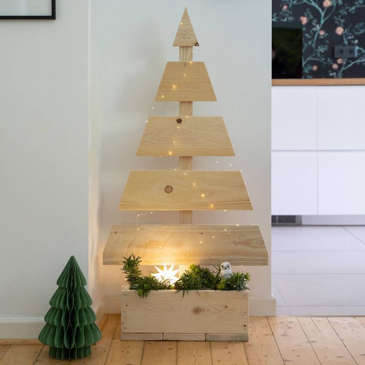 Holzdekoration in Form eines stilisierten Weihnachtsbaums mit Blumen im Kasten auf einem Balkon. Im Hintergrund ist ein Garten mit viel Grün zu sehen.