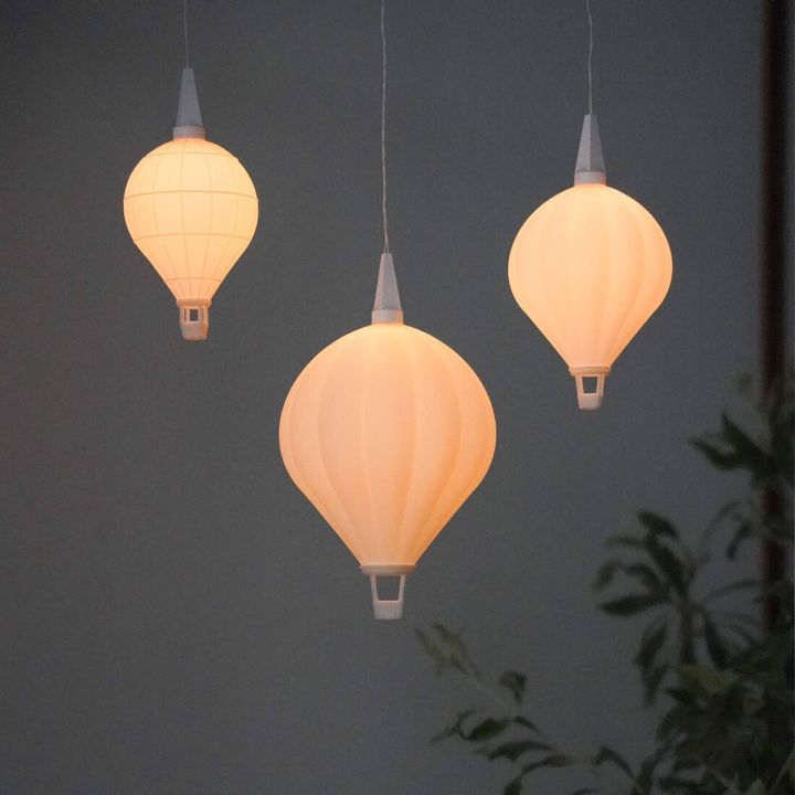 H&auml;ngelampe in Form eines leuchtenden Hei&szlig;luftballons vor grauem Hintergrund. Die Lampe hat eine ballonartige Form aus wei&szlig;em Material mit einer kleinen Korbstruktur am unteren Ende und einer silbernen Aufh&auml;ngung oben.