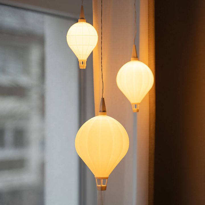 Drei vor einem Fenster h&auml;ngende LED-Lampen in unterschiedlichen Gr&ouml;&szlig;en, geformt wie Hei&szlig;luftballons. Sie haben warmwei&szlig; leuchtende Kunststoffschirme mit Korb unten und Kabelaufh&auml;ngung oben. Die hintere Lampe hat ein abweichendes Rastermuster.