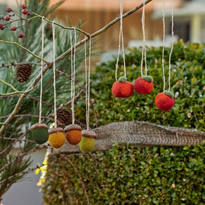 Filz-Dekoanhänger in Form von Eicheln und Äpfeln hängen an einem Zweig über einem Busch. Eicheln sind in Grün, Orange und Gelb gefärbt, Äpfel in Rot und Orange.