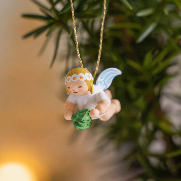 Ein Holzengel schwebt an einem goldenen Faden, hält einen grünen Tannenbaum und hat blaue Flügel. Der Engel trägt ein weißes, gepunktetes Kopftuch. Der Hintergrund ist unscharf mit grünen Pflanzen.