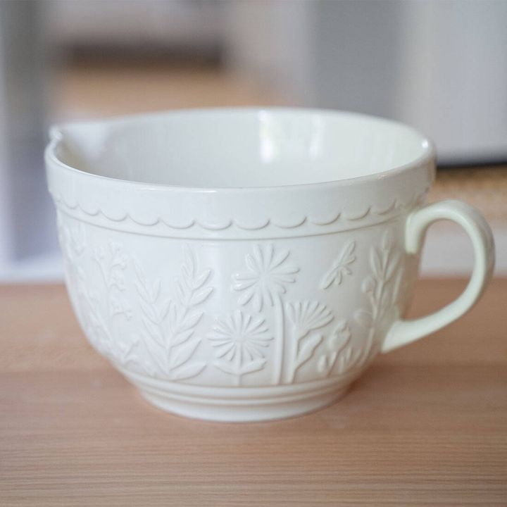 Weiße Keramik-Rührschüssel mit floralem Reliefmuster und einem Ausgießer. Die Schüssel hat einen Henkel und steht auf einer Holztischoberfläche. Der Hintergrund ist unscharf.