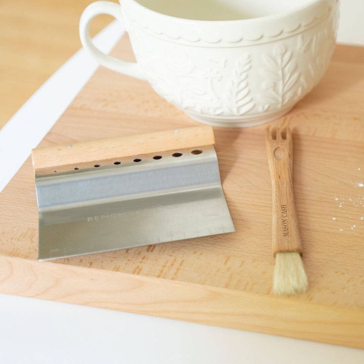 Holzschneidebrett mit einem Metall-Teigschaber und einem Backpinsel aus Holz. Eine große, verzierte, weiße Keramiktasse steht im Hintergrund. Der Teigschaber liegt flach, der Pinsel zeigt zur Bildkante.