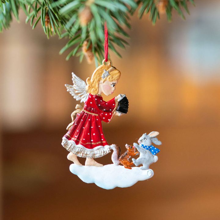 Ein Schmuckanhänger zeigt einen Engel in rotem Kleid mit Harmonika, der auf einer Wolke steht. Vor ihm sitzen Tiere, ein Hase mit blauem Schal, ein Eichhörnchen und ein Vogel. Der Anhänger hängt an einem Tannenzweig.