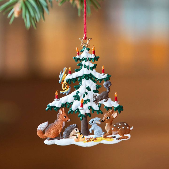 Ein dekorativer Anhänger zeigt einen mit Schnee bedeckten Weihnachtsbaum mit brennenden Kerzen. Unter dem Baum sitzen Tiere: ein Fuchs, ein Igel, ein Hase und ein Reh. Der Anhänger hängt an einem Tannenzweig.