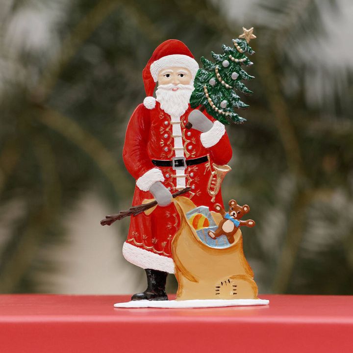 Ein Weihnachtsmann aus Zinn in rotem Anzug hält einen geschmückten Tannenbaum. Neben ihm steht ein Sack voller Spielzeug, darunter ein Teddybär und ein Ball. Die Figur steht auf einem roten Untergrund vor verschwommenem Hintergrund.