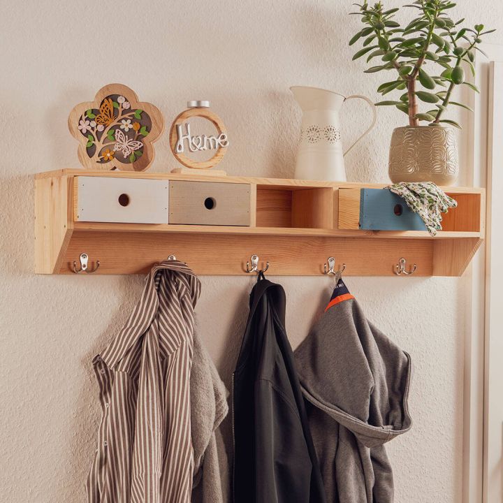 Ein hölzernes Wandregal mit Kleiderhaken darunter. Auf dem Regal stehen Dekorationsgegenstände, darunter ein Keramikkrug, eine Vase mit Pflanze und ein Schild mit "Home". An den Haken hängen Jacken und ein gestreiftes Hemd.