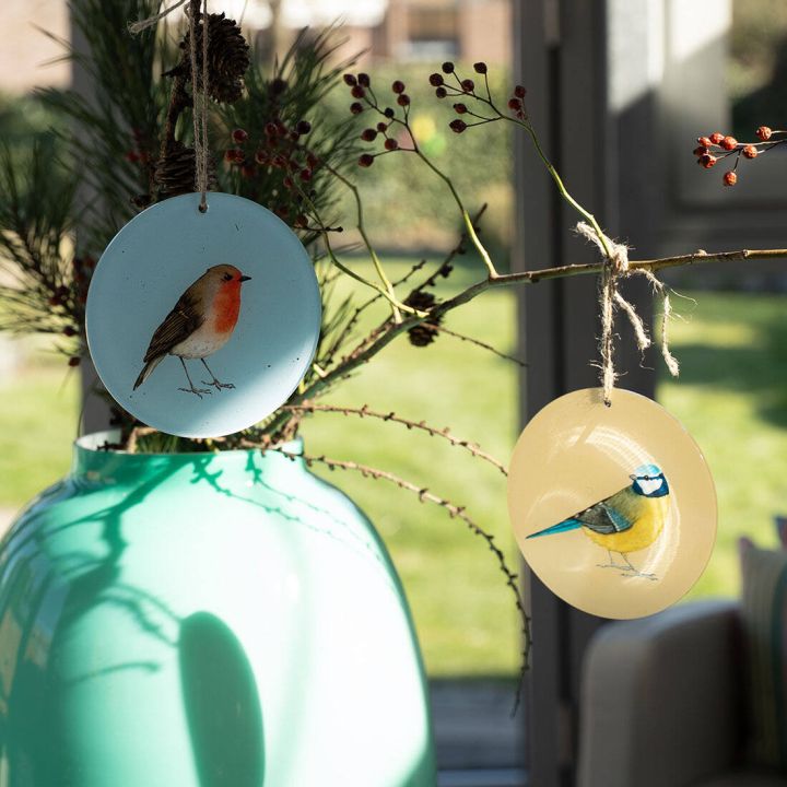 Zwei runde, bemalte Anhänger mit Vogelmotiven hängen an Zweigen in einer großen grünen Vase; der Hintergrund zeigt einen sonnigen Garten.