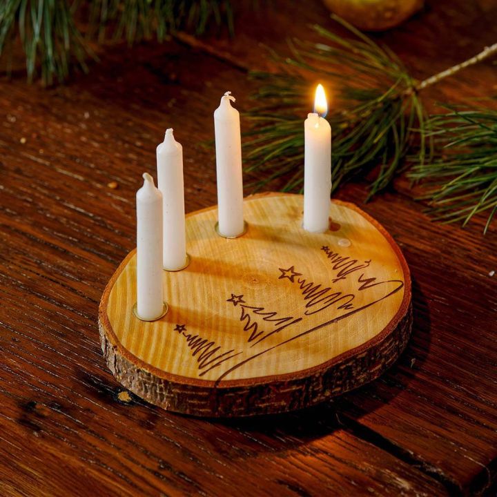 Holzscheibe mit gravierten Tannenbaum-Motiven als Kerzenhalter, best&uuml;ckt mit vier wei&szlig;en, schmalen Kerzen, von denen eine brennt; dekoriert auf einem Holztisch mit Tannenzweigen im Hintergrund.