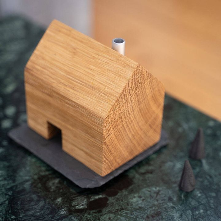 Holzhaus mit rechteckiger Form und Schornstein aus Metall, auf rechteckiger Steinplatte. Platziert auf dunkler Marmoroberfläche, flankiert von zwei kleinen Kegeln, möglicherweise als Baumdeko. Dezente, minimalistische Gestaltung.