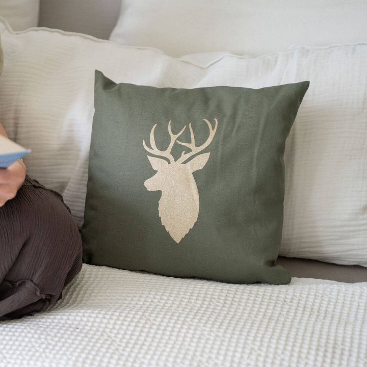 Ein grünes Kissen mit einem beigefarbenen Hirschkopf-Motiv liegt auf einem Bett mit weißen Kissen im Hintergrund. Eine Person sitzt daneben und hält ein Buch.
