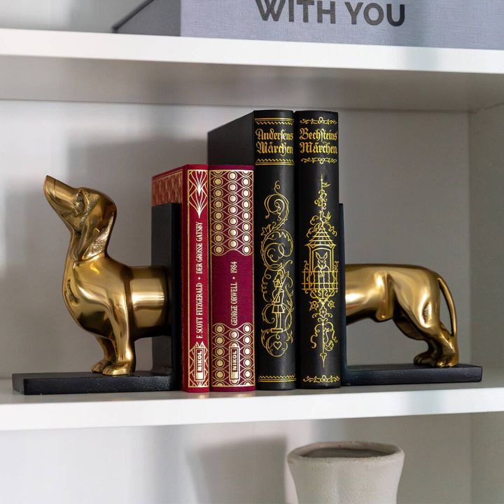 Zweiteiliges Buchst&uuml;tzen-Set in Form eines goldfarbenen Dackels, dessen Vorder- und Hinterteil auf schwarzen Sockeln mehrere dekorative, teils reich verzierte B&uuml;cher auf einem wei&szlig;en Regalbrett aufrecht halten.