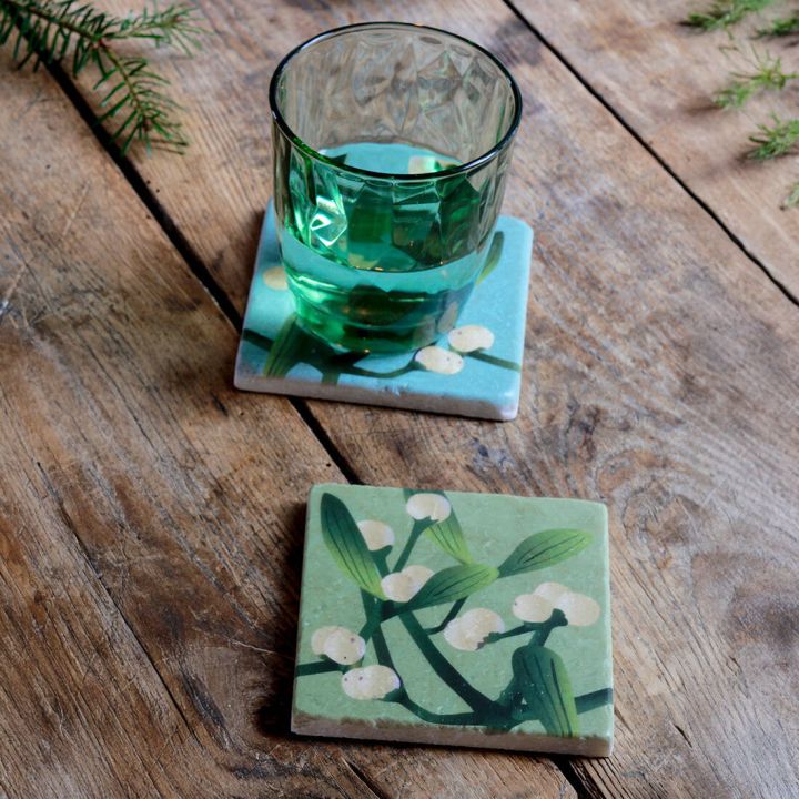 Zwei quadratische Keramikuntersetzer mit gr&uuml;n-blauem Hintergrund und Mistelzweig-Motiv aus gr&uuml;nen Bl&auml;ttern und wei&szlig;en Beeren auf einer rustikalen Holzoberfl&auml;che, einer mit einem gr&uuml;nen Wasserglas, der andere leer im Vordergrund platziert.