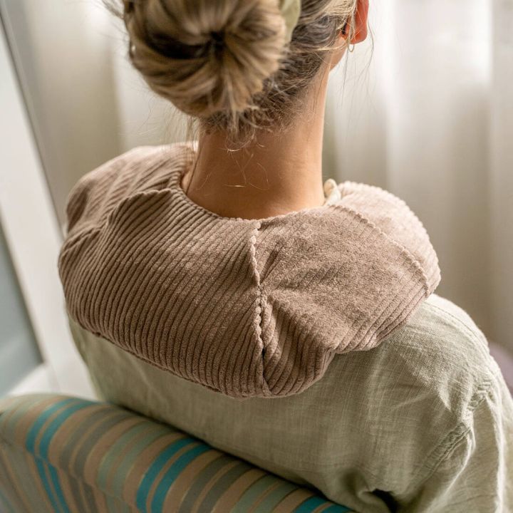 Person von hinten sitzend auf einem Stuhl, tr&auml;gt ein beigefarbenes, geripptes Nacken- und Schulterkissen, das sich anatomisch um Nacken und obere Schultern legt; Fokus auf Passform und Position des W&auml;rmekissens im Einsatz.