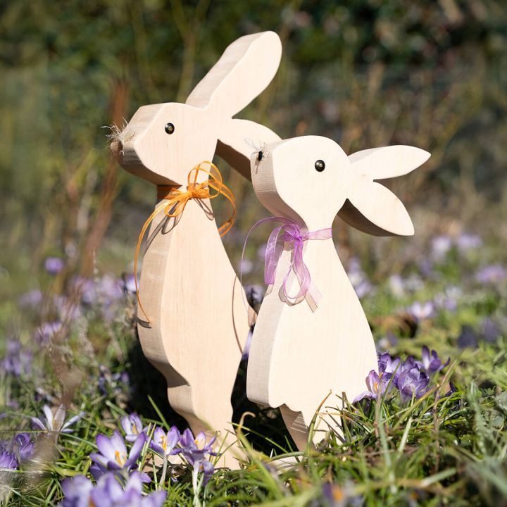 Zwei helle Holzhasen mit bunten Schleifen stehen auf dunkler Erde, umgeben von lila Krokusbl&uuml;ten im Sonnenlicht.