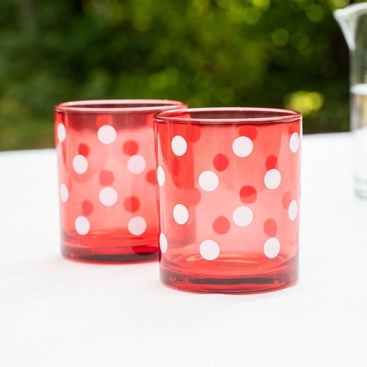 Zwei rote Trinkgl&auml;ser mit wei&szlig;en Punkten stehen auf einem wei&szlig;en Tisch, unscharfer gr&uuml;ner Hintergrund.
