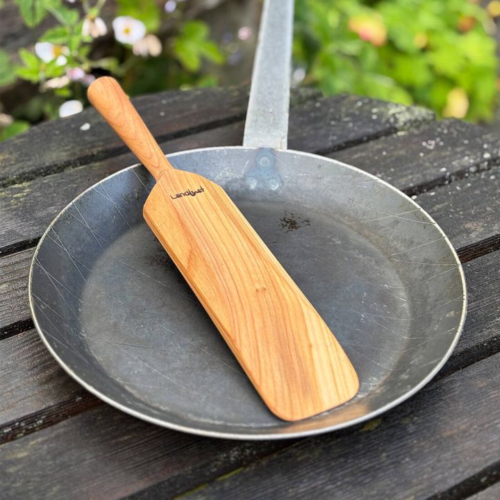 Runde, silberne Pfanne mit Kochpaddel auf dunklem Holztisch, im Hintergrund gr&uuml;ne Pflanzen und Blumen.